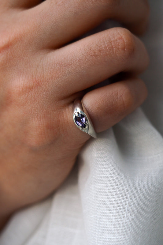 Close-up of a hand wearing a silver ring with a purple gemstone on a light fabric background.