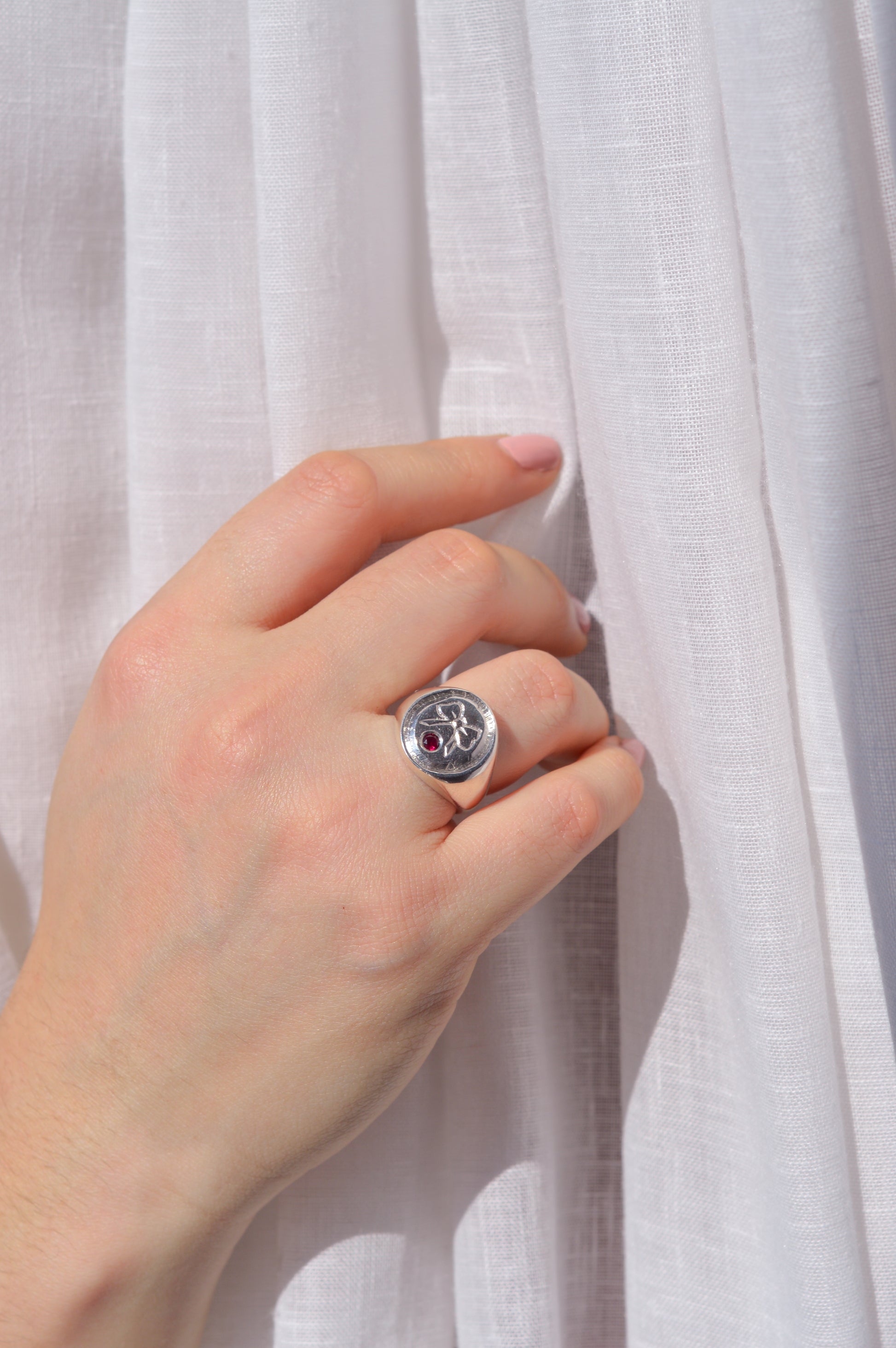 Hand wearing a silver ring with a red gemstone and hand engraved bow against a white fabric background