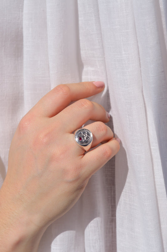 Hand wearing a silver ring with a red gemstone and hand engraved bow against a white fabric background