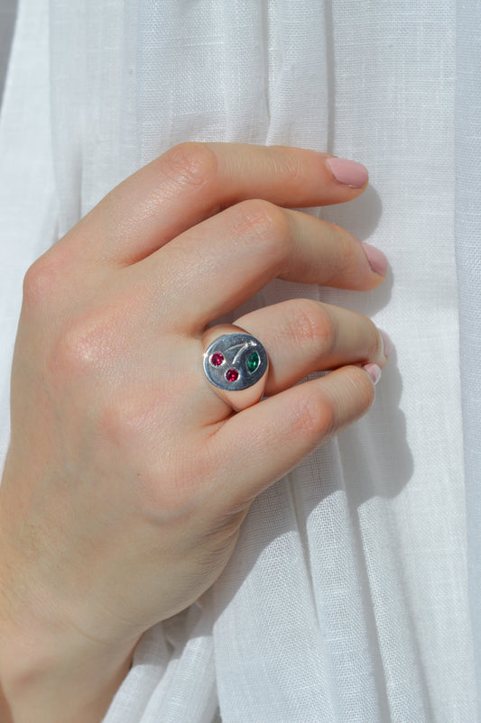 Hand wearing a silver ring with cherry shaped gemstones against a white fabric background
