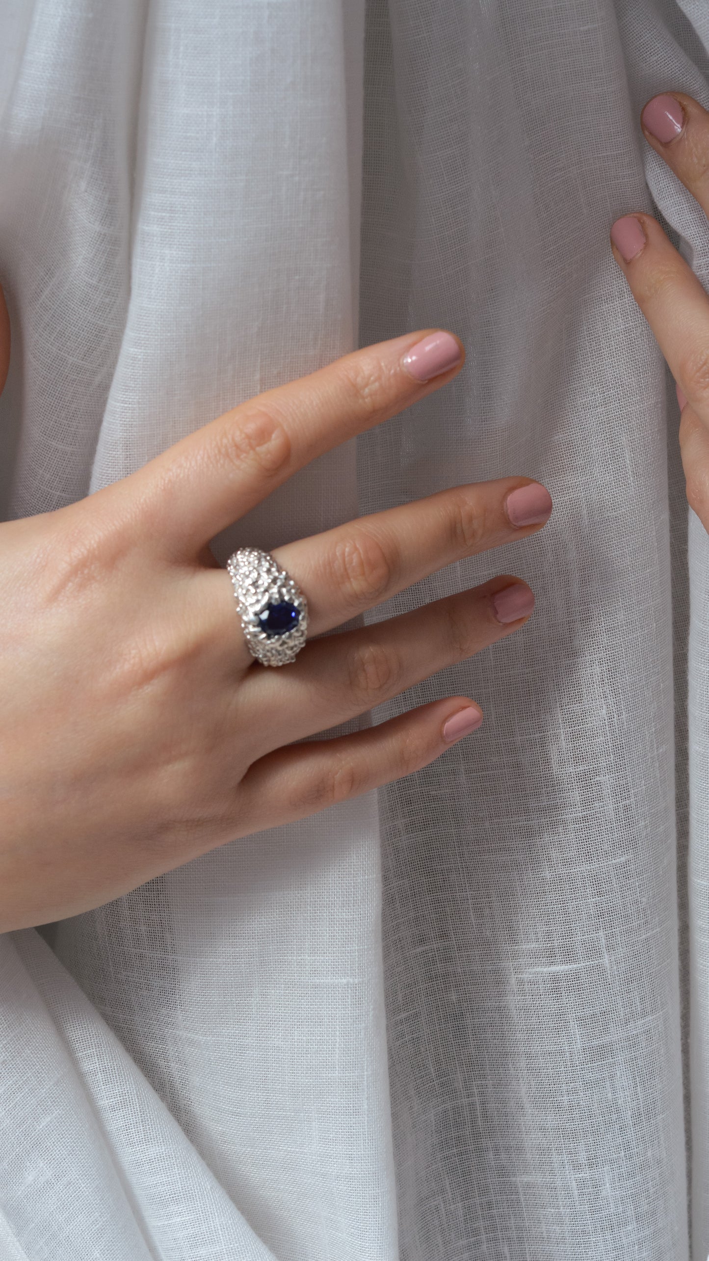 Hand wearing a silver ring with a blue gemstone against a light fabric background