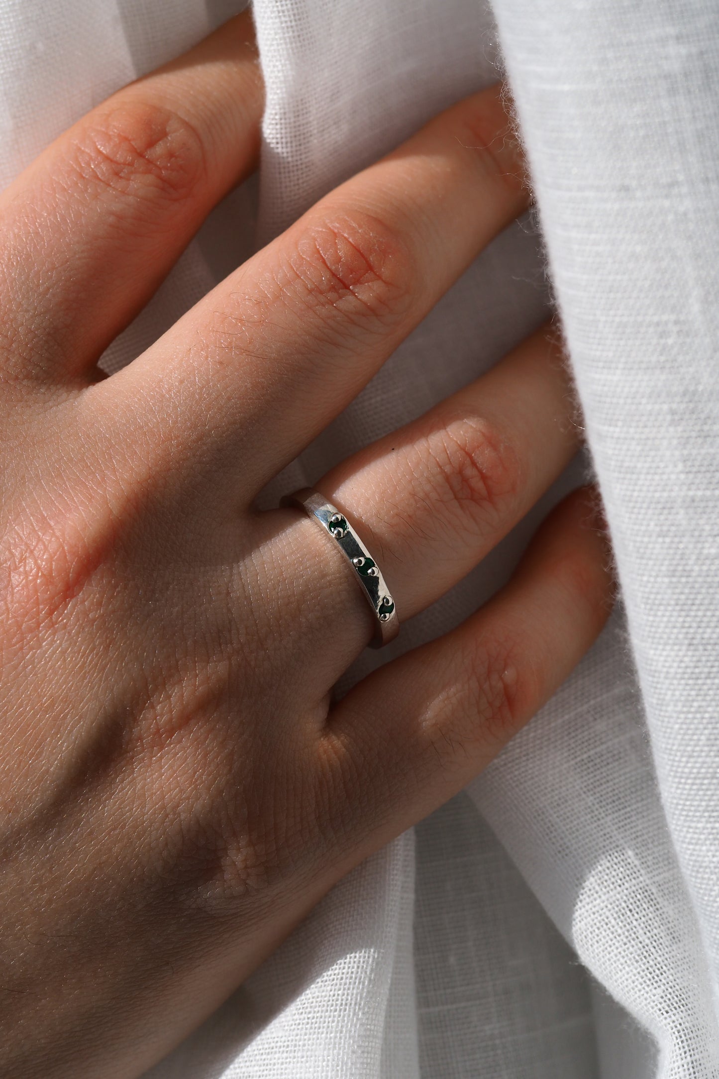 Hand wearing a silver ring with green gemstones against a white fabric background