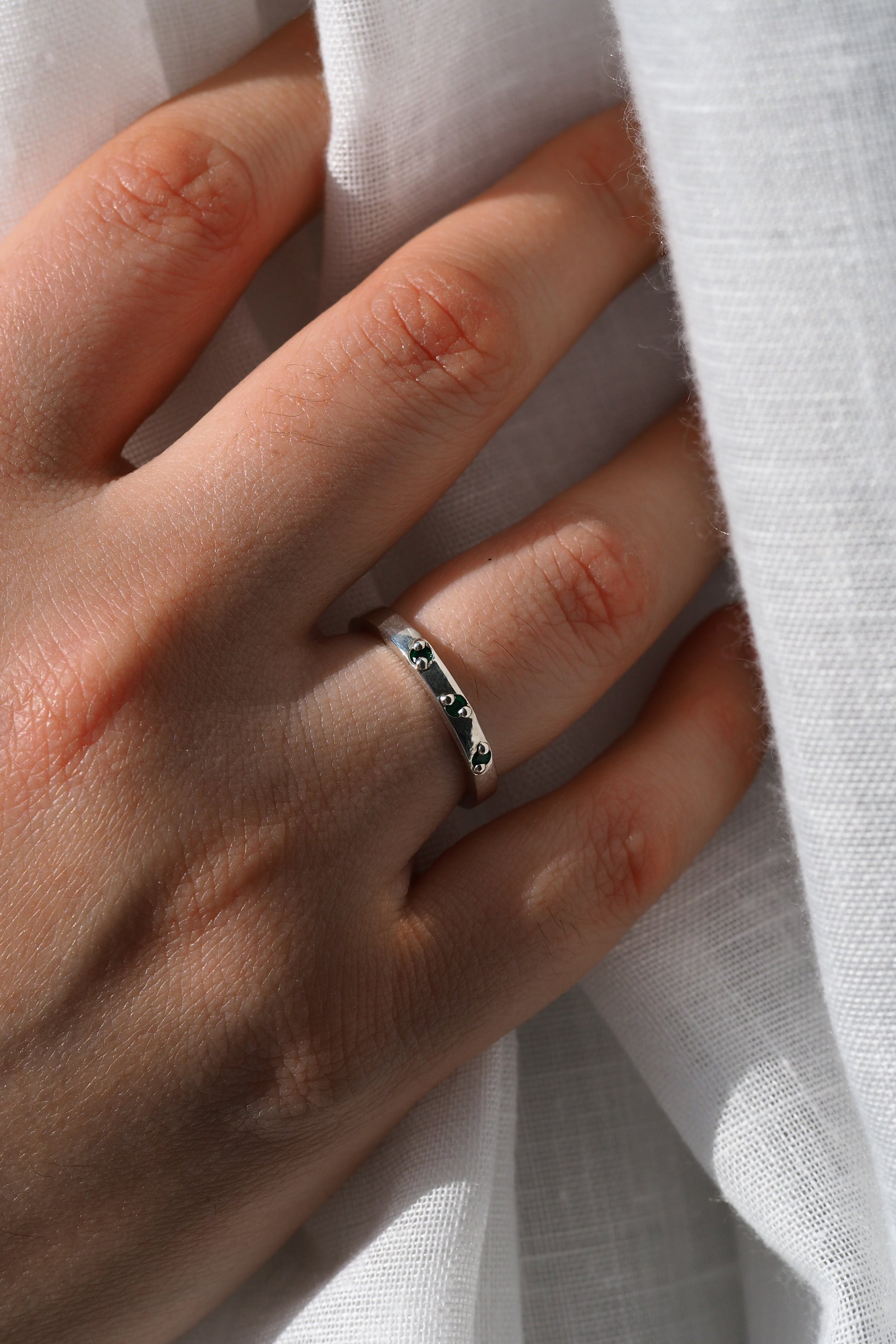 Hand wearing a silver ring with green gemstones against a white fabric background