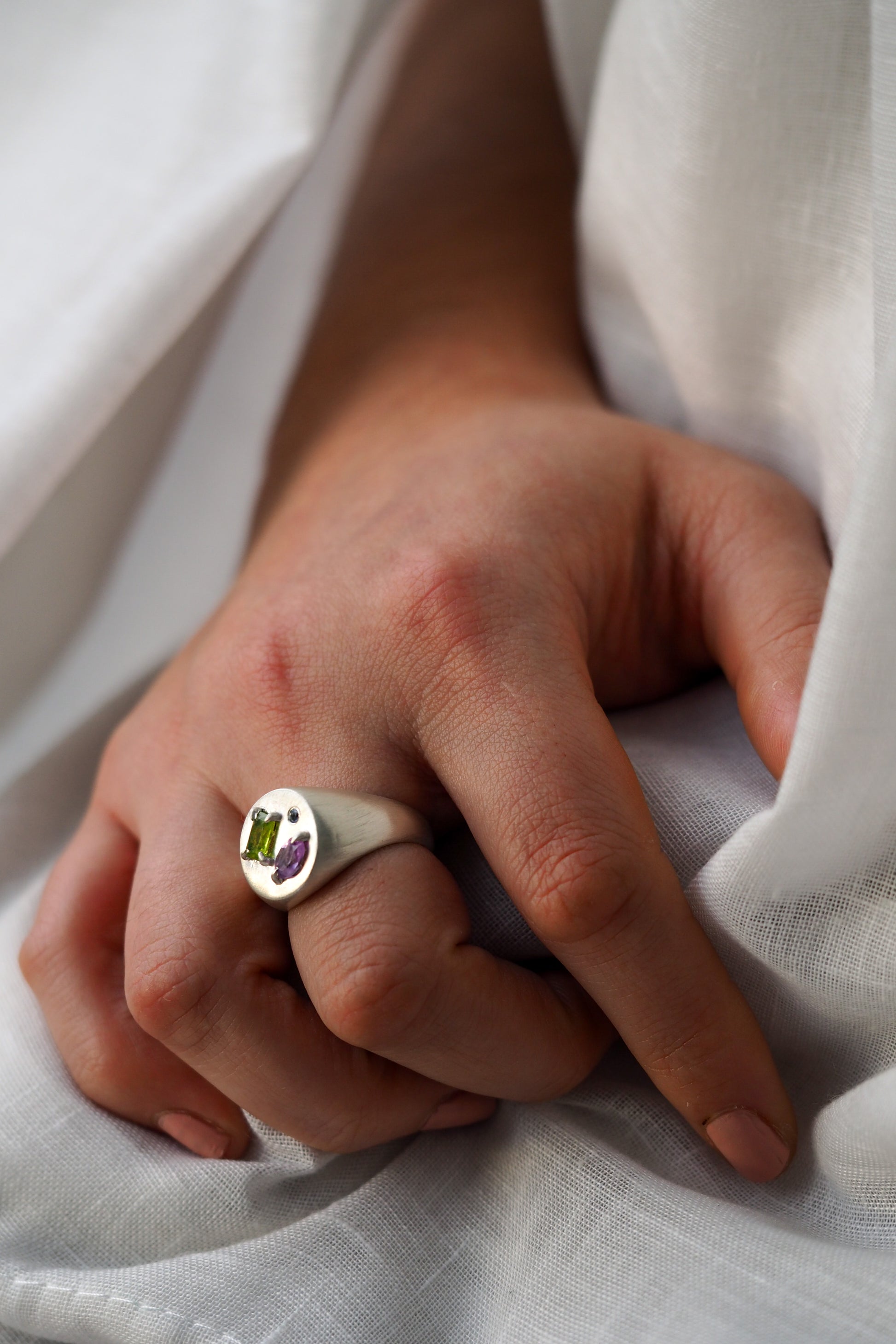 Close-up of a hand wearing a ring with a green gemstone on a white fabric background