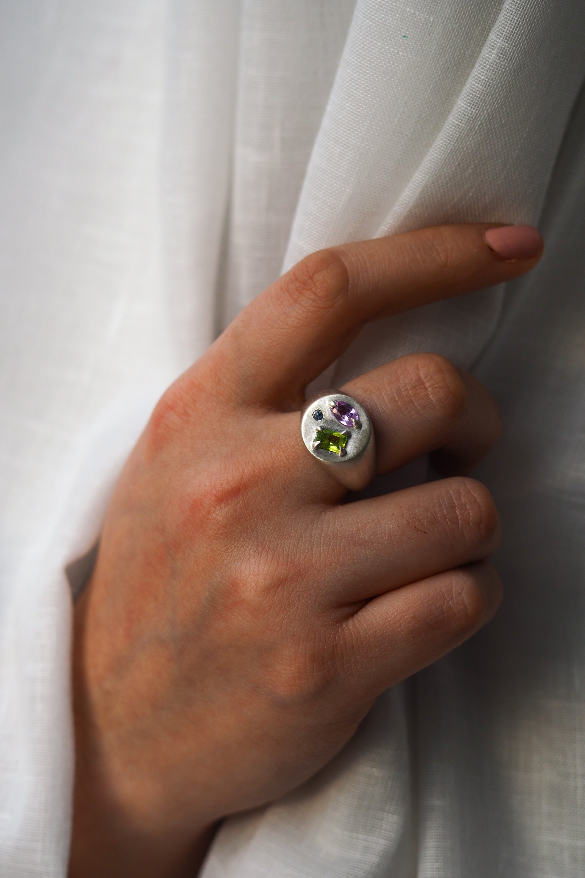 Hand wearing a ring with multicolored gemstones against a white fabric background